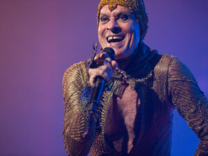 Cantor Ney Matogrosso se apresenta com figurino dourado e brilhante, segurando o microfone e sorrindo durante show, sob luzes azuladas e roxas.