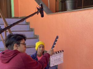 Jovem segura claquete diante de atriz sentada em escada durante gravação de cena em espaço cultural com paredes coloridas em tons de laranja e lilás. Microfone boom aparece suspenso na parte superior da imagem. Cena retrata atividade prática de oficina de cinema.