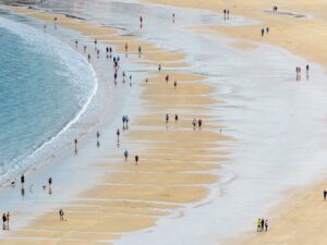 Pessoas caminham ao longo de uma ampla faixa de areia molhada à beira-mar durante a maré baixa, em uma praia movimentada. O mar calmo e as ondas suaves tocam a costa, enquanto grupos e indivíduos aproveitam o passeio.