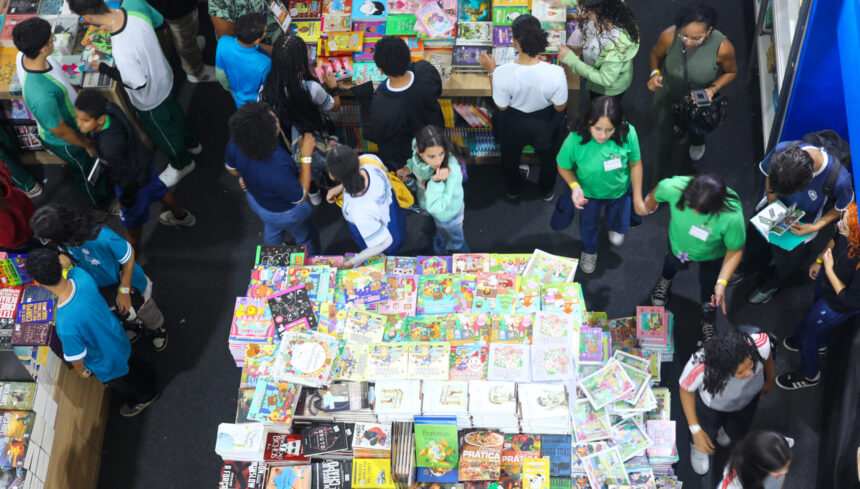 Alunos exploram estandes repletos de livros durante a Bienal Mineira do Livro 2025, em Belo Horizonte. A imagem, capturada de cima, mostra crianças e adolescentes uniformizados folheando obras de diferentes gêneros literários em um ambiente movimentado e colorido.