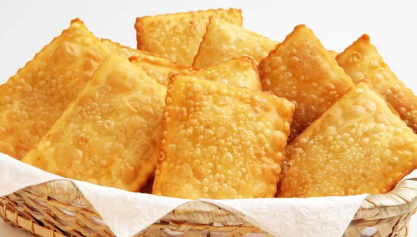 Cesta de vime com vários pastéis fritos retangulares e dourados, sobre papel toalha branco, em fundo branco.