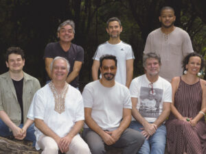 Grupo de dez músicos posa sentado e em pé sobre um tronco de árvore em meio à vegetação do Parque Municipal, em Belo Horizonte. Todos sorriem, vestindo roupas casuais, em clima descontraído. A imagem foi captada durante o dia, com luz natural e árvores ao fundo.