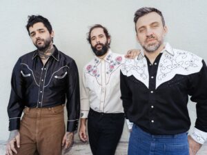 Legenda alt (acessibilidade): Os três integrantes da banda Fresno posam lado a lado em frente a uma parede clara. Da esquerda para a direita: Lucas Silveira, com camisa preta brilhante e calça marrom; Thiago Guerra, usando camisa branca bordada com flores; e Gustavo Mantovani (Vavo), com camisa preta e branca com detalhes rendados e calça jeans.