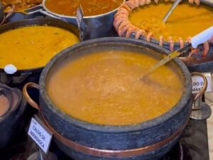 Vista de uma mesa de rodízio de caldos com diversas panelas de pedra e alumínio contendo caldos variados, como caldo de mandioca, frutos do mar, mocotó, pé de galinha e "mexilhão", acompanhados de bandejas com ingredientes e petiscos como pão, calabresa e pimentas.