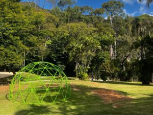 Estrutura metálica em forma de domo, pintada de verde, instalada sobre a grama em um parque arborizado, com céu parcialmente nublado e árvores ao fundo.