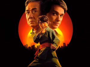 Pôster do filme Karatê Kid: Lendas, com fundo em tons de laranja e vermelho simulando o pôr do sol. Em primeiro plano, o jovem Li Fong (Ben Wang) em pose de luta. Atrás dele, os rostos sérios de Sr. Han (Jackie Chan) e Daniel LaRusso (Ralph Macchio). A silhueta da cidade de Nova York aparece na base da imagem.