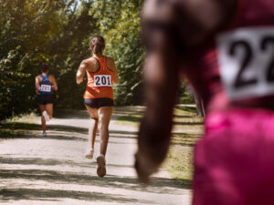 Mulheres correndo em trilha arborizada durante prova de rua, com números de identificação nas costas e sol filtrando pelas árvores.