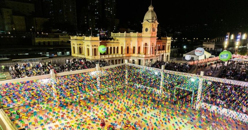 Vista aérea da Praça da Estação, em Belo Horizonte, decorada com bandeirinhas coloridas e arquibancadas lotadas durante o Arraial de Belô à noite.