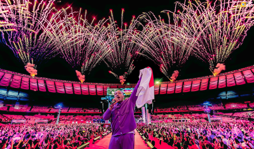 Thiaguinho canta no centro do palco do Mineirão, com camisa nas mãos, diante de uma plateia lotada e fogos de artifício coloridos iluminando o céu ao fundo.