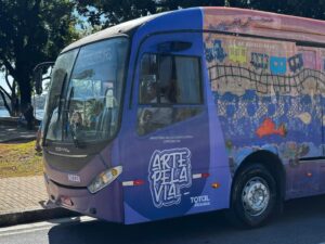 Ônibus decorado com arte urbana do projeto Arte pela Via, com predominância de tons roxos, azuis e elementos gráficos coloridos. Ao fundo, é possível ver a Igreja da Pampulha, em Belo Horizonte