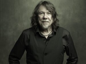 Lenine, de cabelos longos e grisalhos, veste camisa e colete pretos enquanto posa de frente para a câmera, com as mãos nos bolsos, diante de um fundo escuro esverdeado