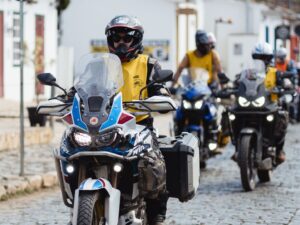 Grupo de motociclistas pilotando por rua de paralelepípedos em Tiradentes, Minas Gerais, durante o Bike Fest. Ao fundo, casas coloniais e público acompanhando o evento