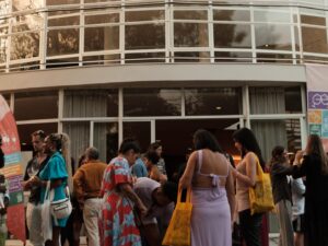 Grupo de pessoas reunidas em frente a prédio moderno durante o festival Vinho na Vila em Belo Horizonte, com banner colorido ao fundo, DJ e saxofonista se apresentando.