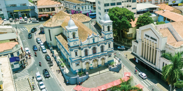 Vista aérea do centro de Nova Lima (MG), mostrando edifícios antigos, ruas movimentadas com carros e pedestres, e arquitetura tradicional da cidade.