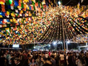 Multidão participa de festa junina à noite, sob um grande teto decorado com bandeirinhas coloridas e luzes, em espaço ao ar livre