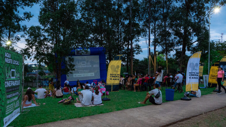 Pessoas reunidas em parque assistindo a filme projetado em tela inflável durante o evento CineParque, cercado por árvores, faixas promocionais e clima de fim de tarde.