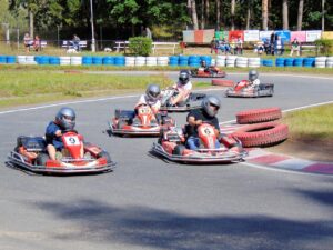 Grupo de pessoas pilotando karts vermelhos em pista externa, usando capacetes; ao fundo, público observa a corrida atrás de barreiras de pneus coloridos.
