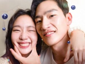 Um homem e uma mulher sul-coreanos estão juntos e sorrindo. O homem encosta a mão na bochecha da mulher de forma carinhosa.