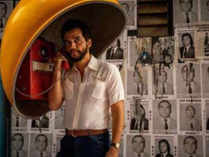 Wagner Moura interpreta Marcelo, um homem moreno vestido com camisa branca e calça preta, atento a uma ligação enquanto está em um orelhão.