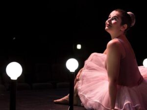 Na foto, uma jovem está sentada olhando para cima com uma expressão serena. Ela está vestida de bailarina, com um traje rosa e o cabelo preso em coque.