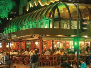 Fotografia noturna de um bar iluminado com várias mesas e pessoas sentadas.