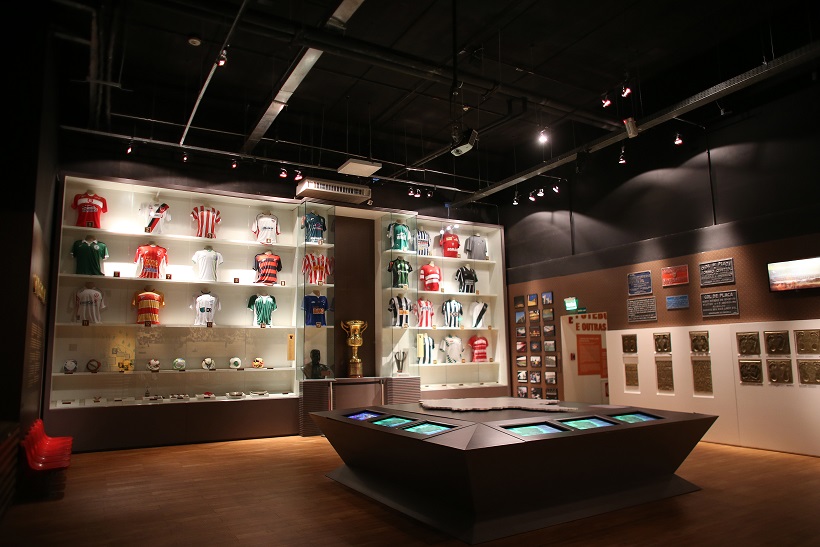 Sala expositiva do Museu Brasileiro do Futebol no Mineirão com camisas históricas de clubes, troféus e mesas interativas ao centro.