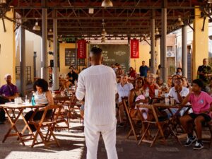 Pessoa se apresenta em um espaço coberto enquanto o público, sentado em mesas de madeira, acompanha atentamente.