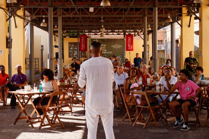 Pessoa se apresenta em um espaço coberto enquanto o público, sentado em mesas de madeira, acompanha atentamente.