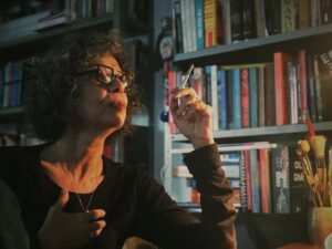 Mulher de roupas escuras segurando um cigarro com a mão, em frente a uma estante de livros.