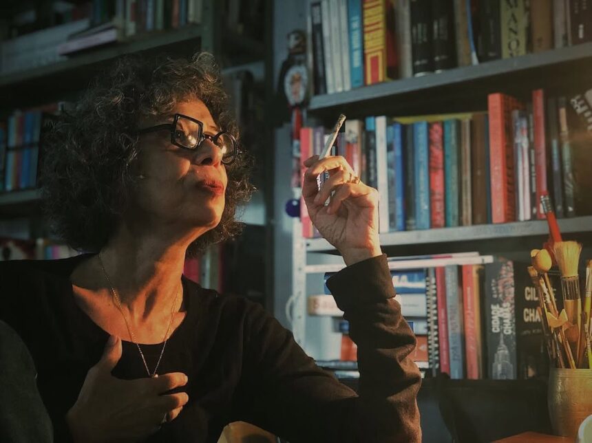 Mulher de roupas escuras segurando um cigarro com a mão, em frente a uma estante de livros.