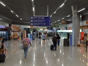 Pessoas caminham pelo saguão do BH Airport, carregando malas e mochilas.