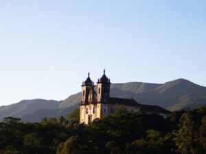 Igreja colonial iluminada pelo sol poente em colina com árvores e montanhas ao fundo.