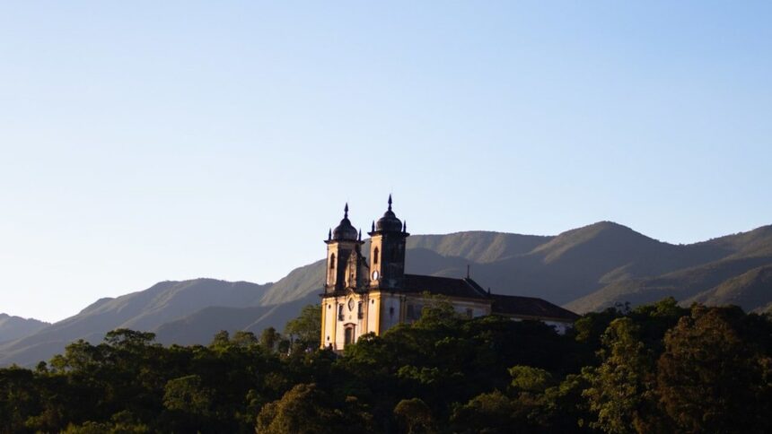 Igreja colonial iluminada pelo sol poente em colina com árvores e montanhas ao fundo.