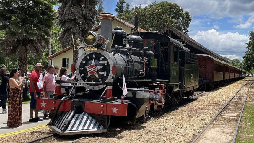 Trem Maria Fumaça preto com número 68 parado em estação, com passageiros e palmeiras.