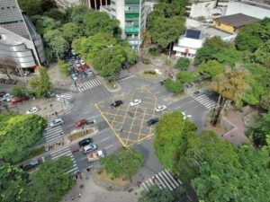 Vista aérea de cruzamento urbano com carros, pedestres, edifícios e muitas árvores verdes.