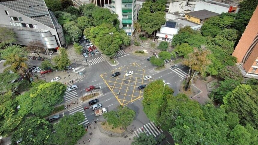 Vista aérea de cruzamento urbano com carros, pedestres, edifícios e muitas árvores verdes.