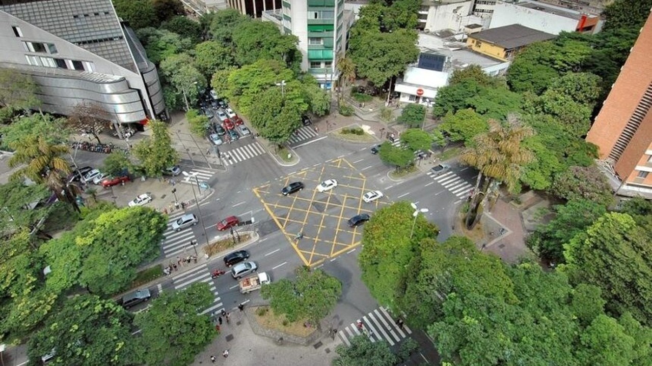 Vista aérea de cruzamento urbano com carros, pedestres, edifícios e muitas árvores verdes.