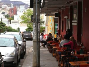Pessoas em bar na calçada. Rua com carros estacionados e edifícios ao fundo.