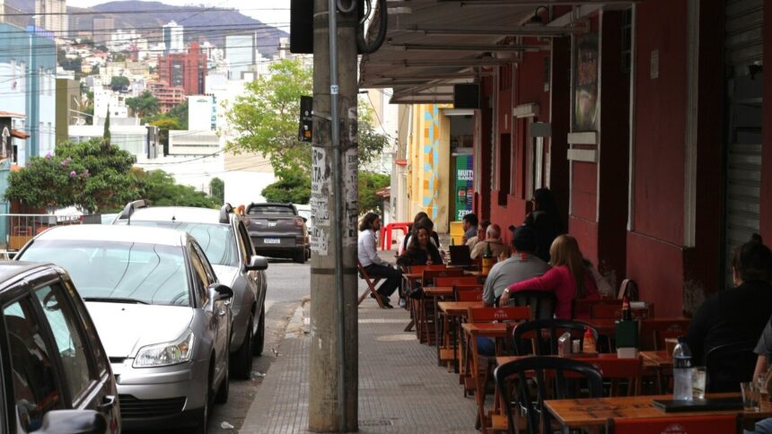 Pessoas em bar na calçada. Rua com carros estacionados e edifícios ao fundo.