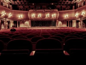 Interior de um teatro com muitas cadeiras de veludo vermelho vazias e um homem sentado em uma delas.