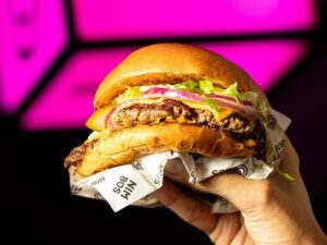 Mão segura hambúrguer duplo com queijo, alface, cebola, picles em papel "NIM BOS" e fundo rosa.