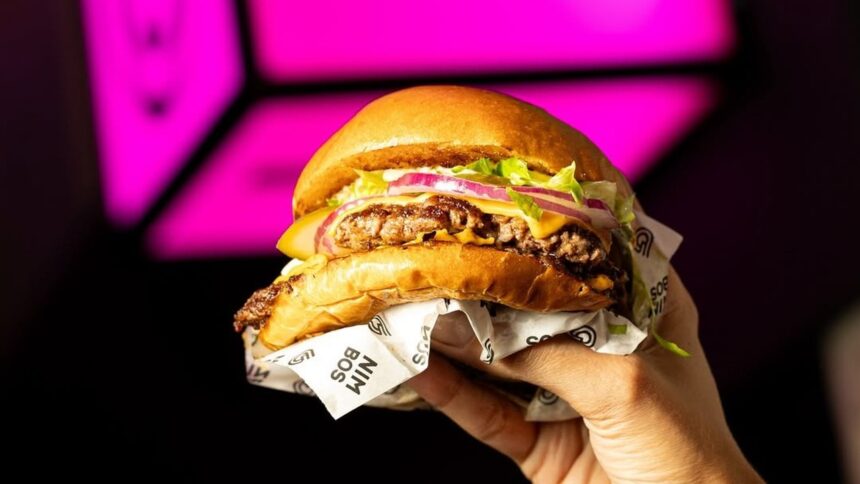 Mão segura hambúrguer duplo com queijo, alface, cebola, picles em papel "NIM BOS" e fundo rosa.