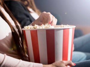 Mãos de mulher pegando pipoca de balde vermelho e branco em cinema escuro.