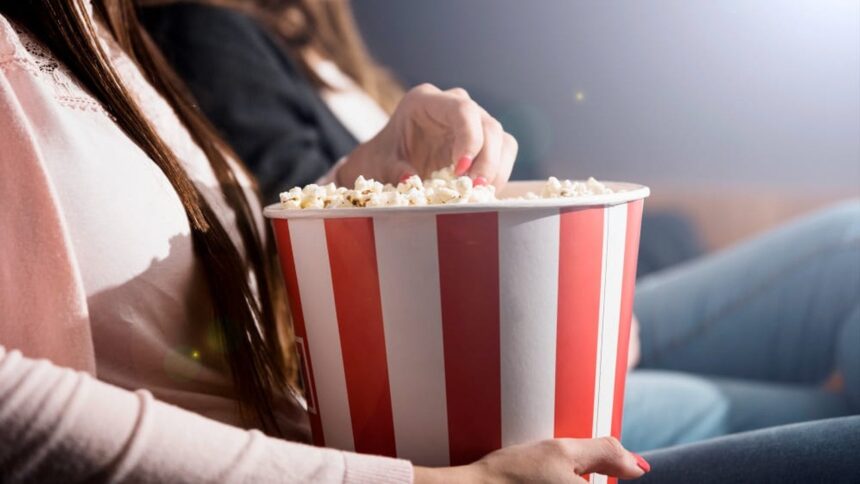 Mãos de mulher pegando pipoca de balde vermelho e branco em cinema escuro.