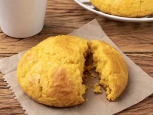 Broa de fubá dourada, partida ao meio, em papel sobre mesa de madeira, com xícara.
