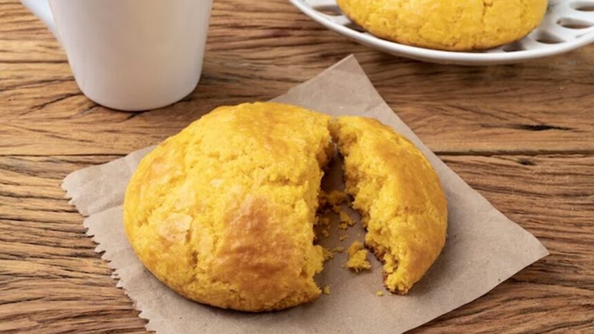 Broa de fubá dourada, partida ao meio, em papel sobre mesa de madeira, com xícara.