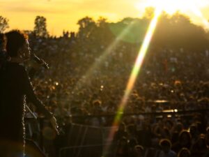 Cantora em silhueta no palco, microfone na mão, performa para grande público em festival ao pôr do sol.
