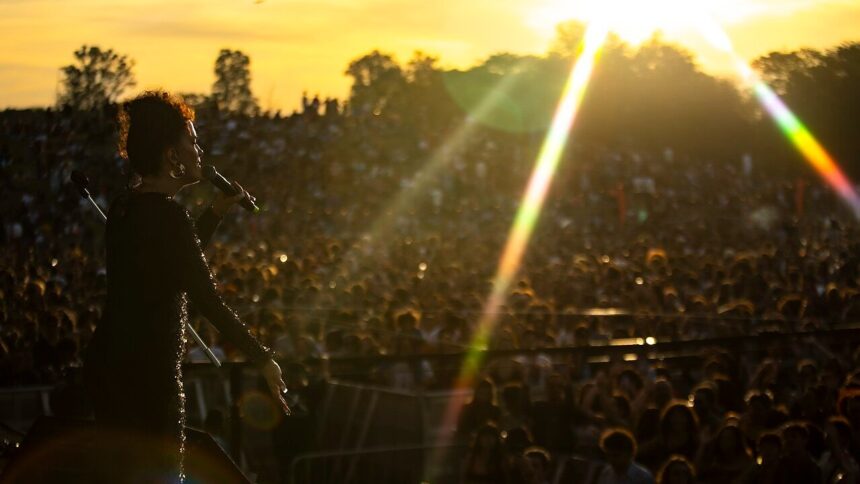 Cantora em silhueta no palco, microfone na mão, performa para grande público em festival ao pôr do sol.