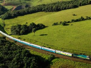 Trem de passageiros azul, cinza e amarelo-esverdeado atravessando uma paisagem rural montanhosa e verde.