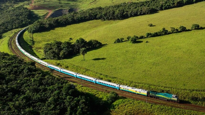 Trem de passageiros azul, cinza e amarelo-esverdeado atravessando uma paisagem rural montanhosa e verde.